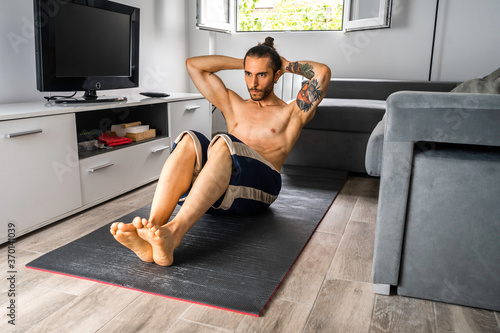 hombre joven en buena forma física, con moño, sin camiseta, y con pantalón corto, haciendo abdominales en una esterilla en el suelo en el salón de su piso de soltero, entrenando