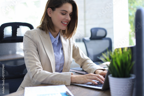 Smiling pretty woman sitting at table, looking at laptop screen. Happy entrepreneur reading message email with good news, chatting with clients online.