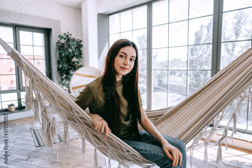 Portrait of attractive young caucasian brunette woman relaxing in the home, sitting at the hammock in light living room with big windows.