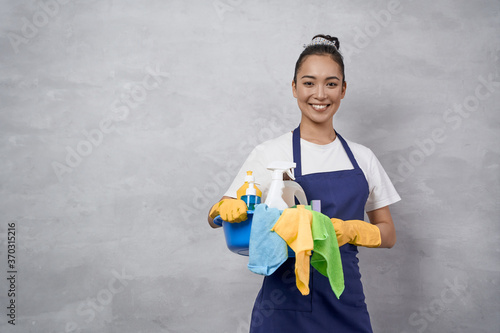 Young happy woman, female cleaner in uniform and rubber gloves holding bucket of cleaning supplies and smiling at camera, standing against grey wall