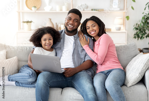 Cheerful Black Family Of Three Relaxing On Couch At Home With Laptop