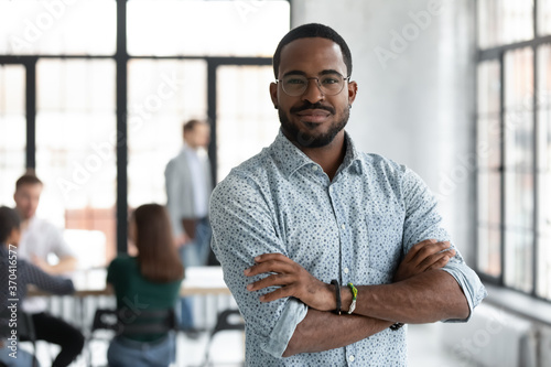 Head shot portrait confident young African American businessman wearing glasses looking at camera, successful executive startup founder standing in modern office room with arms crossed