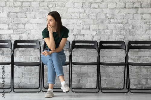 Thoughtful stressed unemployed young woman candidate waiting for job interview or hr manager employer decision, sitting on chair alone in empty office hall, feeling nervous, recruitment concept