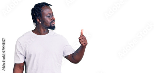 Young african american man with braids wearing casual white tshirt looking proud, smiling doing thumbs up gesture to the side