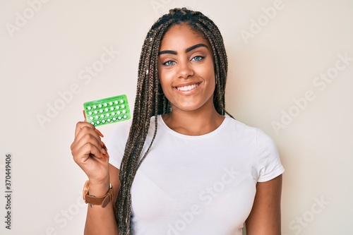 Young african american woman with braids holding birth control pills looking positive and happy standing and smiling with a confident smile showing teeth