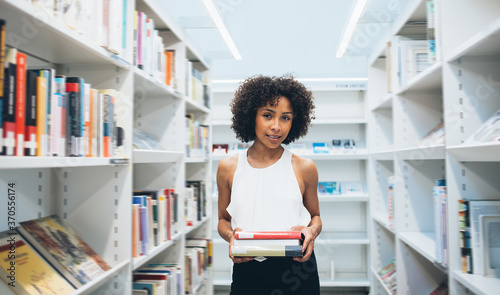 Portrait of attractive dark skinned woman student spending free time in library holding favorite fiction books, beautiful curly african american female looking at camera standing near shelves in store
