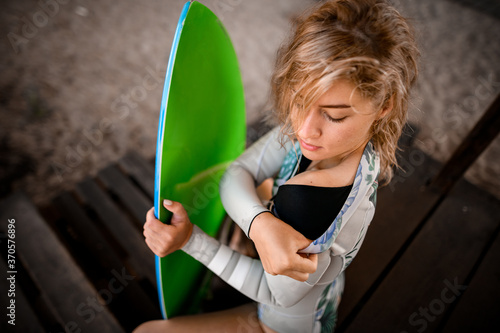 high angle view of young blonde woman which holding surfboard and taking off her wetsuit