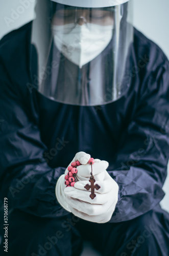 Doctora orando con las manos jutas y un crucifijo durante la pandemia por Covid 19. Mujer con traje de protección, máscara y guantes con una cruz en sus manos mientras ora a dios durante la pandemia.