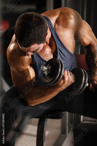 Fitness in gym, sport and healthy lifestyle concept. Handsome athletic man in blue shirt making exercises. Bodybuilder male model training biceps muscles with dumbbell