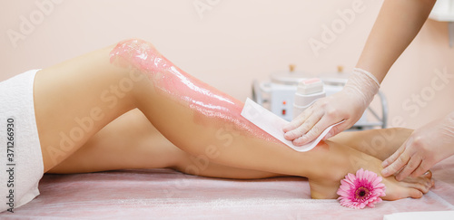 A master applies pink depilatory wax to a young woman's leg for hair removal. Depilation with wax. Beauty concept. Place for text. Selective focus.