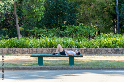 Mulher deitada sobre banco de concreto em parque da cidade de São Paulo, relaxando ouvindo musica com fones de ouvido e celular. 