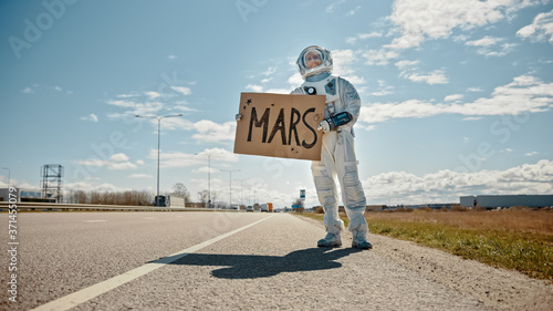 Man in Spacesuit is Standing at the Edge of a Road and Holding a Sign with Mars Written on it. Astronaut Looking to Hitchhike a Car. Spaceman in Futuristic Suit with Technological Panel on His Hand.