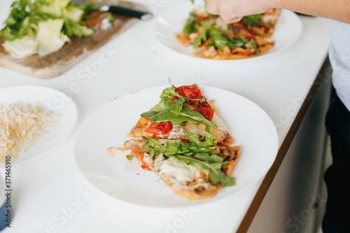 Slice of delicious pizza with mushrooms, tomato, arugula and cheese. Home cooking concept. Hands putting parmesan slices on homemade pizza on plate on modern kitchen.