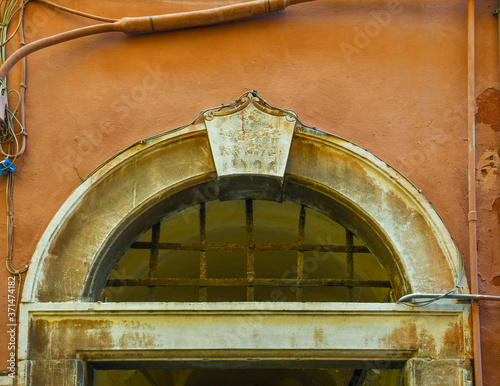 Close-up of the top of the arched stone frame of an old door engraved with the date of 1724 and a rusty metal grate on an orange wall, Italy