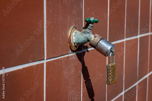  A combination lock on the end of an outdoor water faucet atached to a red grid tiled wall . The image can be a metaphor for drought or water conservation.