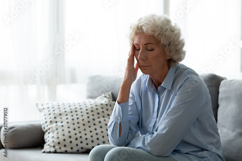 Exhausted mature old hoary woman sitting on sofa, suffering from headache indoors. Nervous middle aged elderly grandmother feeling doubtful about hard decision, health or psychological problems.