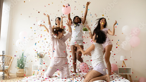 Pajama or bachelorette party celebration concept. Five slim active multiethnic women wearing comfy nightwear sexy glamor pyjamas hanging out jumping dancing on bed under falling multi colored confetti