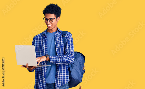 Young african american man holding student backpack using laptop looking positive and happy standing and smiling with a confident smile showing teeth