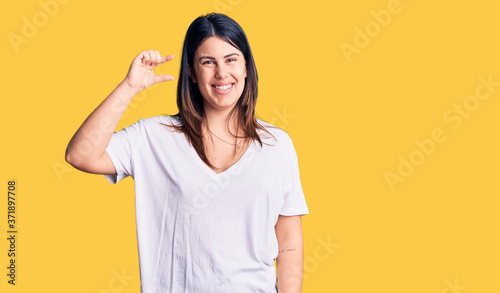 Young beautiful brunette woman wearing casual t-shirt smiling and confident gesturing with hand doing small size sign with fingers looking and the camera. measure concept.