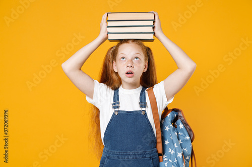 Young fun redhead girl 12-13 years old in white t-shirt backpack hold on head big stack school textbook notebook books isolated on yellow background children studio portrait Kids education concept