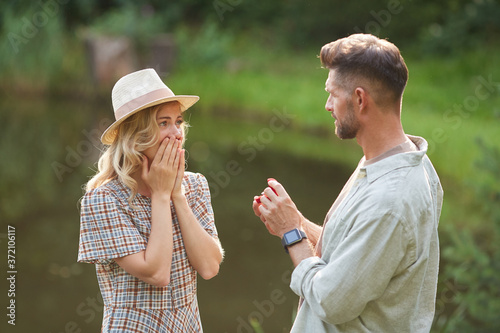 Waist up portrait of man proposing to beautiful girlfriend while standing by lake outdoors in sunlight, copy space