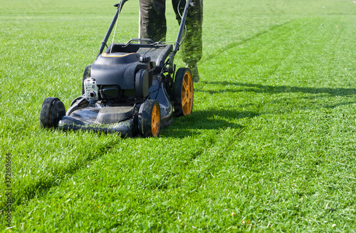 Worker guy shake pour grass from lawn mower bag into wheelbarrow. Garden meadow lawn cutting. Summer works in garden. Static shot.