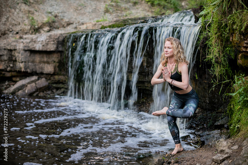 Blonde woman in sportswear practicing yoga at the forest riverside near the waterfall. Fitness girl doing a balance exercise, standing on one leg with hands clasped with in namaste mudra sign
