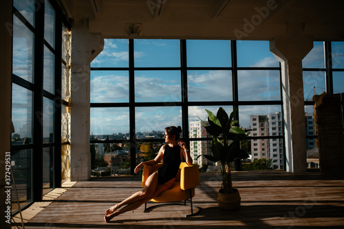 sexy girl with a beautiful figure in a black bodysuit sits on a chair on the background of the window