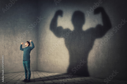 Confident man flexing muscles imagine super power as casting a shadow of muscular bodybuilder showing biceps. Strong person facing his fears. Personal development, inner strength, motivation concept.