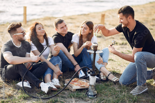 Group of friends is sitting near the lake. They wear casual clothes. Young people eat pizza outside.