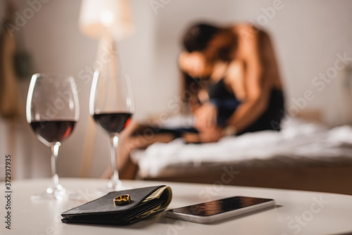 Selective focus of wedding ring on wallet near smartphone and glasses of wine with shirtless man embracing woman on bed