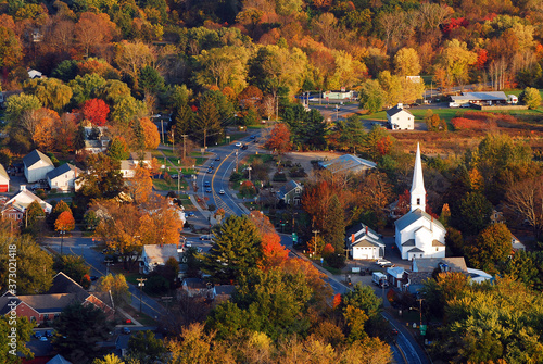 A classic New England town, with a white church with a large steeple, is surrounded by brilliant autumn foliage in an aerial view