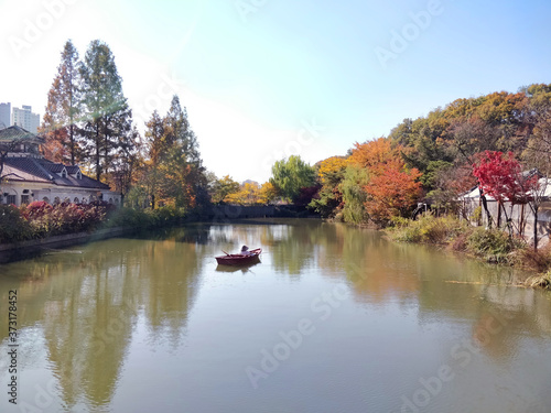 A lonesome boat with a dummy sitting in it floats on a river located inside the Korean Folk Village surrounded by thick fall foliage of various primary colors- Seoul, South Korea- 30th Oct 2018