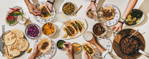 Friends having Mexican Taco dinner. Flat-lay of beef tacos, tomato salsa, tortillas, beer, snacks and peoples hands over white table, top view. Mexican cuisine, gathering, feast, comfort food concept