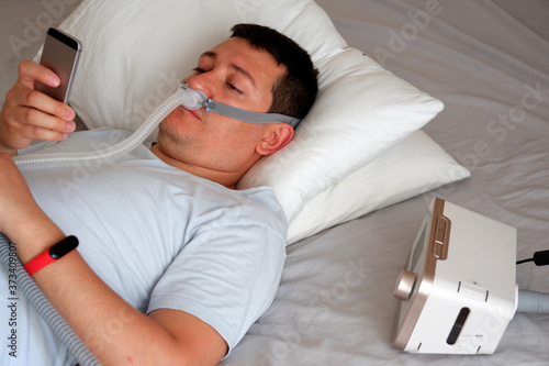 young man wearing under the nose nasal mask and using CPAP machine for sleeping smoothly and playing with his smart phone before sleeping. selective focus.