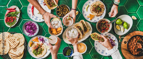 Friends home taco party. Flat-lay of Mexican traditional dishes Tacos with beef meat, corn tortillas , tomato salsa and peoples hands with glasses over green background, top view. Mexican cuisine