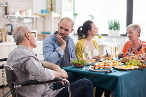 Disabled senior in wheelchair man having a conversation with son during family brunch in kitchen. Senior parents together with mature children.