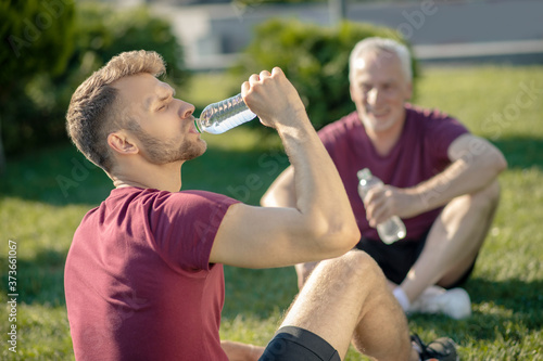 Bearded male drinking water, grey-haired man holding bottle, both sitting on grass