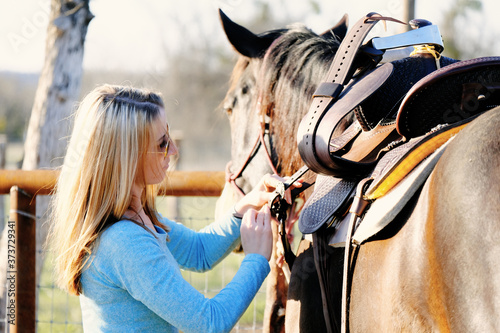 Western lifestyle shows woman putting on saddle to go horseback riding outdoors.