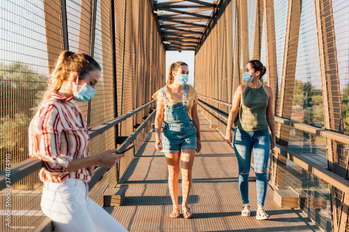 One young woman with medical mask and cellphone and two women with surgical mask walking and talking. New Normal Coronavirus pandemic concept. Young women with face mask outdoor.