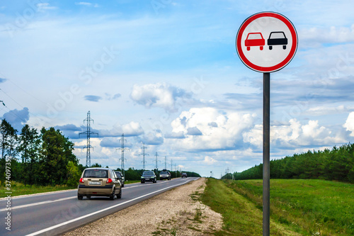 International road sigh 'No overtaking' standing near highway. Fast moving cars on the road. Forest & high voltage towers are on background