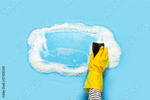 Hand in a yellow rubber glove holds a cleaning sponge and wipes a soapy foam on a blue background. Cleaning concept, cleaning service. Banner. Flat lay, top view