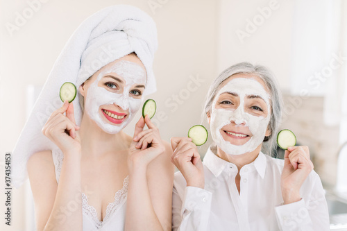 Morning skin care routine, family spa day. Pretty senior gray haired woman and her young attractive granddaughter making clay facial masks and posing to camera in kitchen with cucumber slices