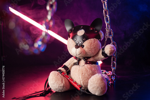 toy bear dressed in leather belts harness accessory for BDSM games on a dark background in neon light