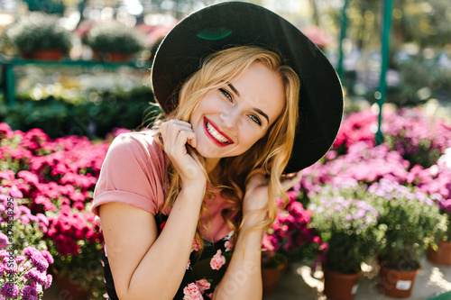 Ecstatic caucasian girl chilling in orangery in morning. Inspired young lady in black hat posing with pink flowers on background