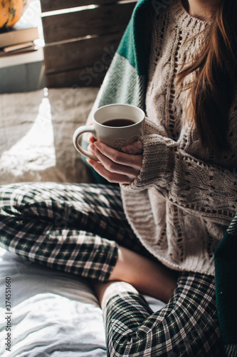 Cozy autumn or winter atmosphere. Young woman with cup of tea and warm sweater is seating at home near the window. Scandinavian hygge concept.