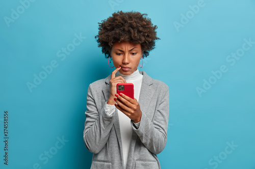 Puzzled confused dark skinned woman in formal wear looks indignant at smartphone, gazes at display, reads business news on website, isolated blue background. People and modern technology concept