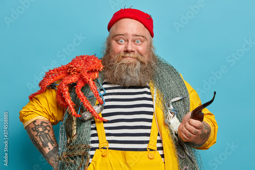 Photo of bearded experienced sailor poses with fishing net, big red crab on shoulders, smoke pipe, welcomes on board, enjoys marine cruise during summer vacation. Bearded captain has sea life