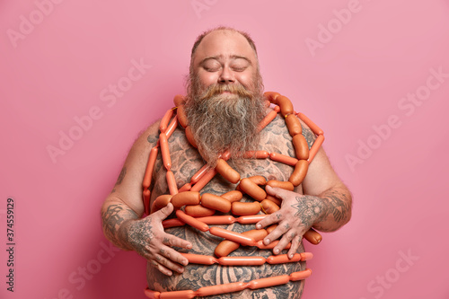 Obese overweight man has unhealthy nutrition, bad habits, wrapped with sausages, has diet failure, feels satiety after eating junk food, regularly overeats, keeps hands on big fat tattooed tummy.