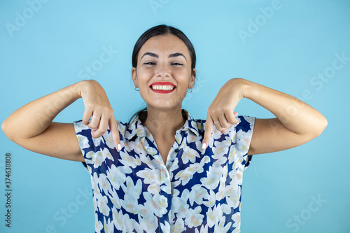 Young beautiful woman wearing sunglasses over isolated blue background smiling, looking and pointing down with fingers and raised arms.